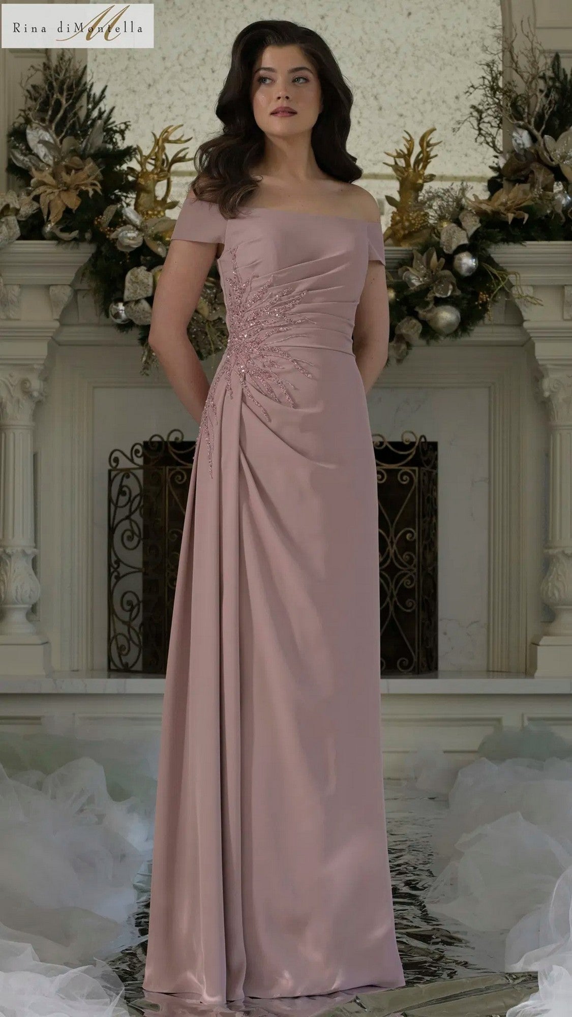 A woman stands indoors in a blush Rina Di Montella RD2806 long formal dress, an off-the-shoulder crepe column gown with ruched detailing, in front of a decorated white mantelpiece with floral arrangements.